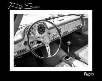 Op de afbeelding: Zwart-witfoto van het interieur van een klassieke auto. Het stuurwiel, het dashboard en de versnellingspook zijn zichtbaar. Het dashboard heeft een zilveren afwerking en de tekst "300 SL" is zichtbaar.