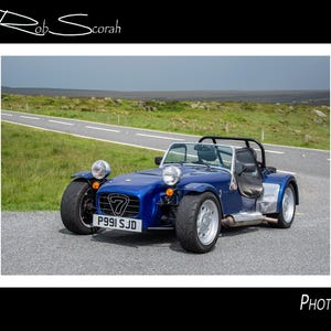 Puede incluir: Un coche deportivo azul con una barra antivuelco negra está estacionado en una carretera. El coche tiene molduras plateadas, neumáticos grandes y una matrícula P991 SJD. El fondo presenta un paisaje verde y un cielo nublado.