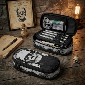 Peut inclure: Une trousse rectangulaire noire et blanche ornée d'une illustration du monstre de Frankenstein. La trousse est ouverte, révélant des boucles élastiques internes contenant plusieurs stylos et une poche en filet avec un câble blanc enroulé.