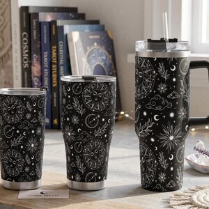 May include: Three black stainless steel tumblers with a white celestial pattern featuring suns, moons, stars, clouds, and foliage. The tumblers have clear lids, and the largest one includes a black handle and a clear straw for drinking.