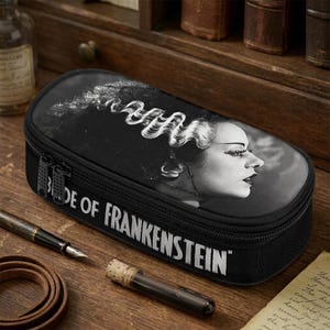 Può includere: Astuccio nero con un'immagine in bianco e nero della Sposa di Frankenstein. L'astuccio ha una chiusura a cerniera e il testo "Bride of Frankenstein" stampato sul davanti.