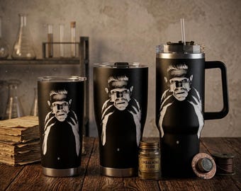 May include: Black insulated tumblers and a mug featuring a white and black graphic of Frankenstein's monster. A small jar labeled "Embalming Fluid" and a spool of wire are also present. The items are arranged on a wooden surface.