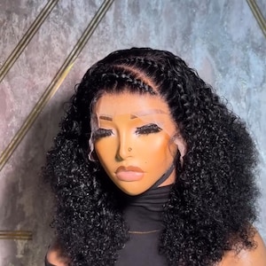 Puede incluir: Una peluca negra rizada de cabello humano con un frente de encaje y cabello trenzado a lo largo de la línea del cabello. La peluca está diseñada en un bob corto con rizos sueltos.