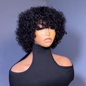 Op de afbeelding: Een zwarte krullende pruik met een pony op een mannequinhoofd. De pruik is gestyled in een korte, stompe bob met strakke krullen. De mannequin draagt een zwart shirt met een hoge hals en uitgesneden schouders.
