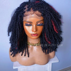 Peut inclure: Une perruque de cheveux humains bouclés noire et rouge avec une dentelle frontale. La perruque est coiffée en un bob court et dégradé avec des mèches rouges. La perruque est sur une tête de mannequin et est posée sur un tabouret en bois blanc. Le texte "THE BEAUTY BAR" est visible dans le coin inférieur droit de l'image.