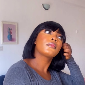 Puede incluir: Una mujer con piel marrón oscura lleva una camisa gris de manga larga. Tiene el pelo negro con flequillo y mira hacia arriba y hacia la derecha. Su maquillaje está hecho con una sombra de ojos marrón claro y un labio brillante.