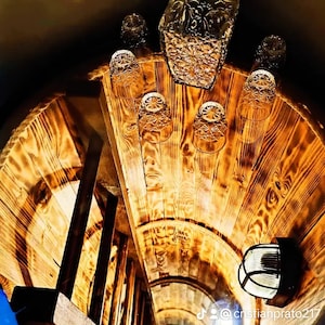 Puede incluir: Un barril de madera con una escalera en su interior. El barril está lleno de vasos de chupito de vidrio transparente y una jarra de vidrio transparente con un tapón. La jarra y los vasos de chupito están decorados con un patrón floral. El barril está iluminado por una pequeña lámpara.