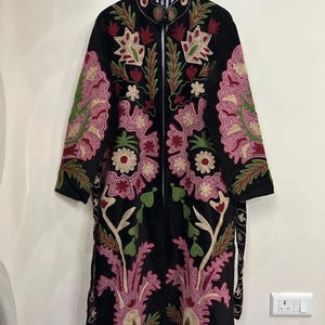 May include: A black long-sleeve coat featuring intricate floral embroidery in shades of pink, red, and green. The garment has a high collar, a front zipper closure, and a visible black and white striped lining along the inner edges of the coat.