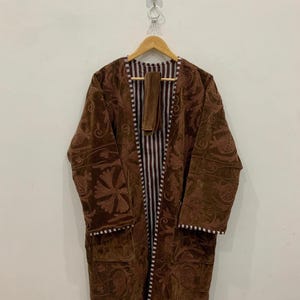 Op de afbeelding: Een lange, bruine badjas met ingewikkelde bloemenborduursels en een bijpassende riem. De badjas heeft een gestreepte voering en witte biezen. Hij hangt aan een houten kleerhanger tegen een effen witte muur.