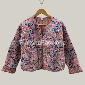 May include: A pink quilted jacket with a floral pattern in shades of pink, blue, and green. The jacket has a V-neck, long sleeves, and a striped trim on the cuffs and collar. The jacket is hanging on a wooden hanger.