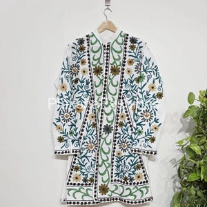 Pode incluir: Casaco branco bordado com um design floral. O casaco apresenta gola mandarim, mangas compridas e fecho de botões. O bordado inclui flores e folhas verdes, castanhas e amarelas. O casaco está pendurado num cabide de madeira.