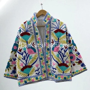 May include: White jacket with colorful floral embroidery. The jacket features a stand-up collar and three-quarter sleeves. The embroidery includes blue, pink, yellow, and red flowers and leaves. The jacket is hanging on a wooden hanger.