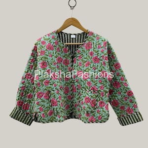 Può includere: Giacca trapuntata con motivo floreale di fiori rosa su sfondo azzurro. La giacca ha un colletto rotondo e maniche lunghe con polsini a righe. La fodera interna è a righe. La giacca è appesa a una gruccia di legno.