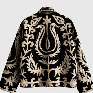 Puede incluir: Una chaqueta negra con intrincados bordados de color crema en la espalda y las mangas. El diseño muestra motivos florales y de hojas simétricos con un patrón de planta estilizada en el centro. La chaqueta tiene cuello alto y mangas largas.