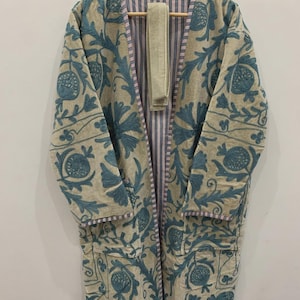 May include: Beige embroidered coat with a blue floral pattern. The coat has long sleeves and a matching belt. The interior lining features a striped pattern. The coat is hanging on a wooden hanger.