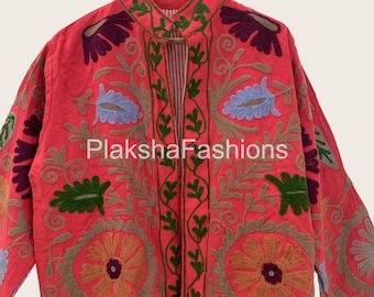 May include: A coral-colored jacket with intricate floral embroidery in shades of green, gold, purple, and blue. The jacket features a mandarin collar and is open in the front. The text "Plaksha Fashions" is visible on the front of the jacket.