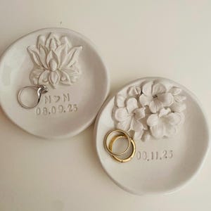 Puede incluir: Dos platos blancos de cerámica para anillos. Uno presenta un diseño de flor de loto, el otro un grupo de flores. Cada plato contiene anillos y tiene una fecha inscrita. El primer plato tiene la inscripción "N>N 08.09.25". El segundo plato tiene la inscripción "00.11.25".