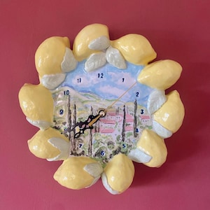 May include: A decorative wall clock featuring a lemon design. The clock face depicts a landscape scene with a blue sky, trees, and buildings. The clock is surrounded by yellow lemons and green leaves. The clock hands are black.
