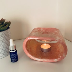 Può includere: Un diffusore di oli essenziali in ceramica rosa e bianca con una candela a lumino accesa all'interno. Una piccola bottiglia blu con un contagocce è accanto al diffusore. Sullo sfondo c'è una pianta grassa in vaso.