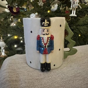 May include: A white ceramic mug with a green tree-shaped handle and a decorative nutcracker figure. The nutcracker is dressed in a blue jacket, red accents, and black boots. Black polka dots are scattered across the mug's surface. Christmas themed.