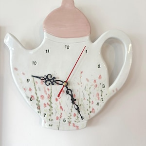Può includere: Un orologio da parete a forma di teiera in ceramica bianca con coperchio e manico rosa. Il quadrante dell'orologio presenta numeri neri, lancette nere ornate e una lancetta dei secondi rossa. Motivi floreali rosa e verdi decorano il quadrante.