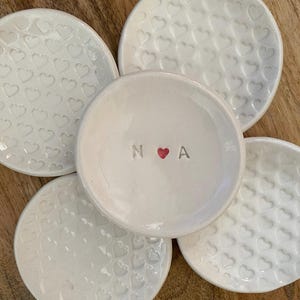 Puede incluir: Un conjunto de pequeños cuencos de cerámica blanca. Cuatro cuencos presentan un patrón de corazones en relieve, mientras que el cuenco central tiene las iniciales "N A" con un corazón rojo. Los cuencos están dispuestos sobre una superficie de madera.