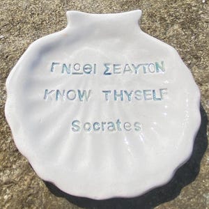 Può includere: Un pezzo di ceramica bianca a forma di conchiglia con l'iscrizione "ΓΝΩΘΙ ΣΕΑΥΤΟΝ, KNOW THYSELF, Socrate" in lettere azzurre. I bordi della conchiglia sono smerlati e la superficie ha una finitura leggermente lucida. Il testo è centrato.