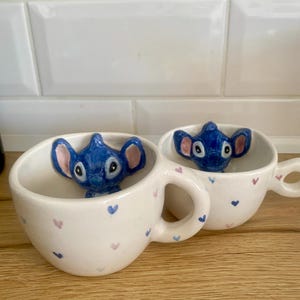 May include: Two white ceramic mugs with a blue Stitch figure inside. The mugs have a heart pattern in pink and blue. The mugs are on a wooden surface.