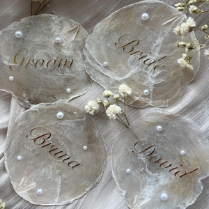 Könnte beinhalten: Vier Platzkarten in Muschelform mit goldfarbenen Buchstaben und Perlenakzenten. Die Karten sind mit "Groom", "Bride", "Bruna" und "David" beschriftet. Weiße Blumen sind um die Karten platziert. Die Karten liegen auf einer hellen, strukturierten Oberfläche.