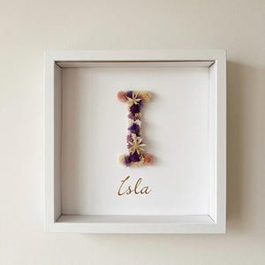 Könnte beinhalten: Ein quadratischer weißer Schattenfugenrahmen mit einem großen Buchstaben I, der aus getrockneten violetten, cremefarbenen und rosafarbenen Blumen besteht. Unter dem Buchstaben ist der Name Isla in goldener Schreibschrift auf dem weißen Hintergrund.