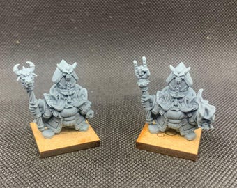May include: Two gray miniature figurines of fantasy characters, each standing on a small wooden base. The figures are detailed with armor, helmets, and staffs. One figure is holding up a hand in a rock and roll gesture.