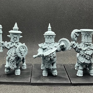Op de afbeelding: Drie grijze miniatuurfiguren van hars die centaur-achtige krijgers met baarden voorstellen, gekleed in versierde helmen en harnassen met schedelmotieven. Elk figuur staat op een vierkante voet en houdt bijlen en ronde schilden met stieremblemen.