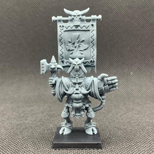 Könnte beinhalten: Graue Miniaturfigur eines Fantasy-Kriegers, der einen Hammer und ein Banner hält. Das Banner zeigt ein Blitzdesign. Der Krieger hat einen langen Bart, einen gehörnten Helm und steht auf einem schwarzen Sockel.