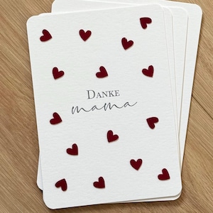 Könnte beinhalten: Ein Stapel weißer Karten mit roten Herzformen, die über die Oberfläche verstreut sind. Auf den Karten steht in einer Schriftart "Danke mama". Die Karten liegen auf einer Holzoberfläche.