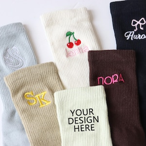 Könnte beinhalten: Sechs Paar Socken mit verschiedenen Stickereien. Die Socken sind in verschiedenen Farben, darunter Weiß, Beige, Hellblau, Braun und Schwarz. Die Designs beinhalten eine Kirsche, eine Schleife, Initialen und die Wörter "Jenna", "Nora" und "Aurora". Ein Paar Socken hat den Text "Your Design Here".