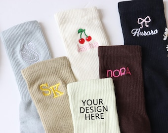 Puede incluir: Seis pares de calcetines con diferentes diseños bordados. Los calcetines son de varios colores, incluyendo blanco, beige, azul claro, marrón y negro. Los diseños incluyen una cereza, un lazo, iniciales y las palabras "Jenna", "Nora" y "Aurora". Un par de calcetines tiene el texto "Your Design Here".