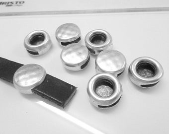 Könnte beinhalten: Silberfarbene Metallperlen mit flacher Oberseite und zylindrischem Körper. Ein schwarzer Lederriemen wird mit einer silbernen Perle gezeigt. Die Perlen sind auf einer weißen Oberfläche verstreut, was auf Schmuckherstellung oder Bastelbedarf hindeutet.