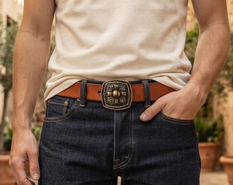 May include: A tan leather belt featuring a square-shaped brass buckle with an intricate embossed pattern and a central circular stud. The belt is worn through the belt loops of dark blue denim jeans, paired with a plain white short-sleeved t-shirt.
