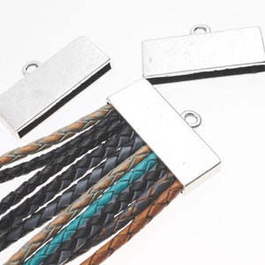 Puede incluir: Tapas de extremo de metal plateadas con un bucle para sujetar a un cordón de cuero. Las tapas de extremo son rectangulares y tienen un acabado liso y pulido. El cordón de cuero está trenzado y viene en una variedad de colores, incluyendo marrón, negro y azul verdoso.