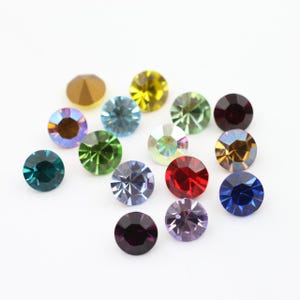 Puede incluir: Surtido de piedras de imitación redondas y facetadas en varios colores, incluyendo verde, azul, rojo, morado y dorado. Las piedras de imitación están dispuestas sobre una superficie blanca.