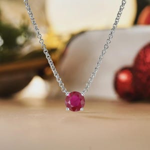 Pode incluir: Um colar de prata com um pendente de gema vermelha intensa e redonda. A corrente é fina, e a gema vermelha é o elemento central. O colar é exibido sobre um fundo desfocado, realçando a elegância da joia.