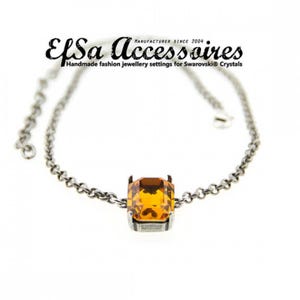 Puede incluir: Una pulsera de cadena plateada con un gran cristal cuadrado de color ámbar como punto focal. El cristal está engastado en un marco de metal plateado. La pulsera tiene el texto "EfSa Accessoires" y "Handmade fashion jewellery settings for Swarovski® Crystals".