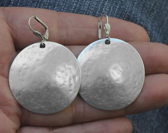 May include: A pair of silver-toned, hammered-metal disc earrings. Each earring is a large, flat circle with a textured surface and a lever-back closure. The earrings are displayed against a neutral background.