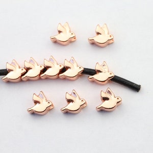 May include: A set of rose gold-colored metal dove charms. The doves are in flight and have a delicate, detailed design. The charms are strung on a black cord.