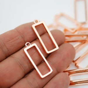 May include: Rose gold rectangular pendant charms with a small loop at the top for attaching to jewelry. The charms are hollow in the center and have a smooth, polished finish. Several charms are visible.