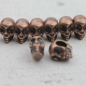 Puede incluir: Primer plano de varias cuentas de calavera de color bronce. Las cuentas están ensartadas juntas, con algunas cuentas sueltas esparcidas por delante. Las calaveras tienen cuencas de ojos grandes y rasgos faciales detallados.