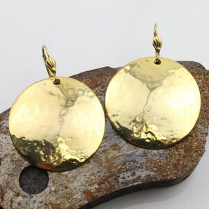 Può includere: Un paio di orecchini a disco in metallo dorato martellato. Ogni orecchino presenta un grande pendente circolare con una superficie strutturata e una chiusura a gancio decorativa. Gli orecchini sono esposti su una superficie marrone ruvida.