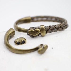 May include: A brown leather bracelet with a heart-shaped metal clasp. The bracelet is made of two strands of leather that are braided together. The clasp is made of metal and has a heart-shaped design.