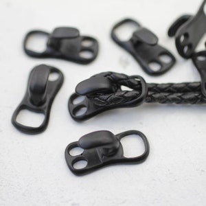 May include: Black metal clasps with two holes and a hook. The clasps are used for attaching straps or cords to clothing or accessories.