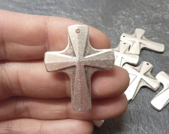 May include: A silver-toned metal cross pendant with a matte, textured finish and a small circular hole at the top for hanging. The cross features a flared design with beveled edges, and several identical pendants are visible in the background.